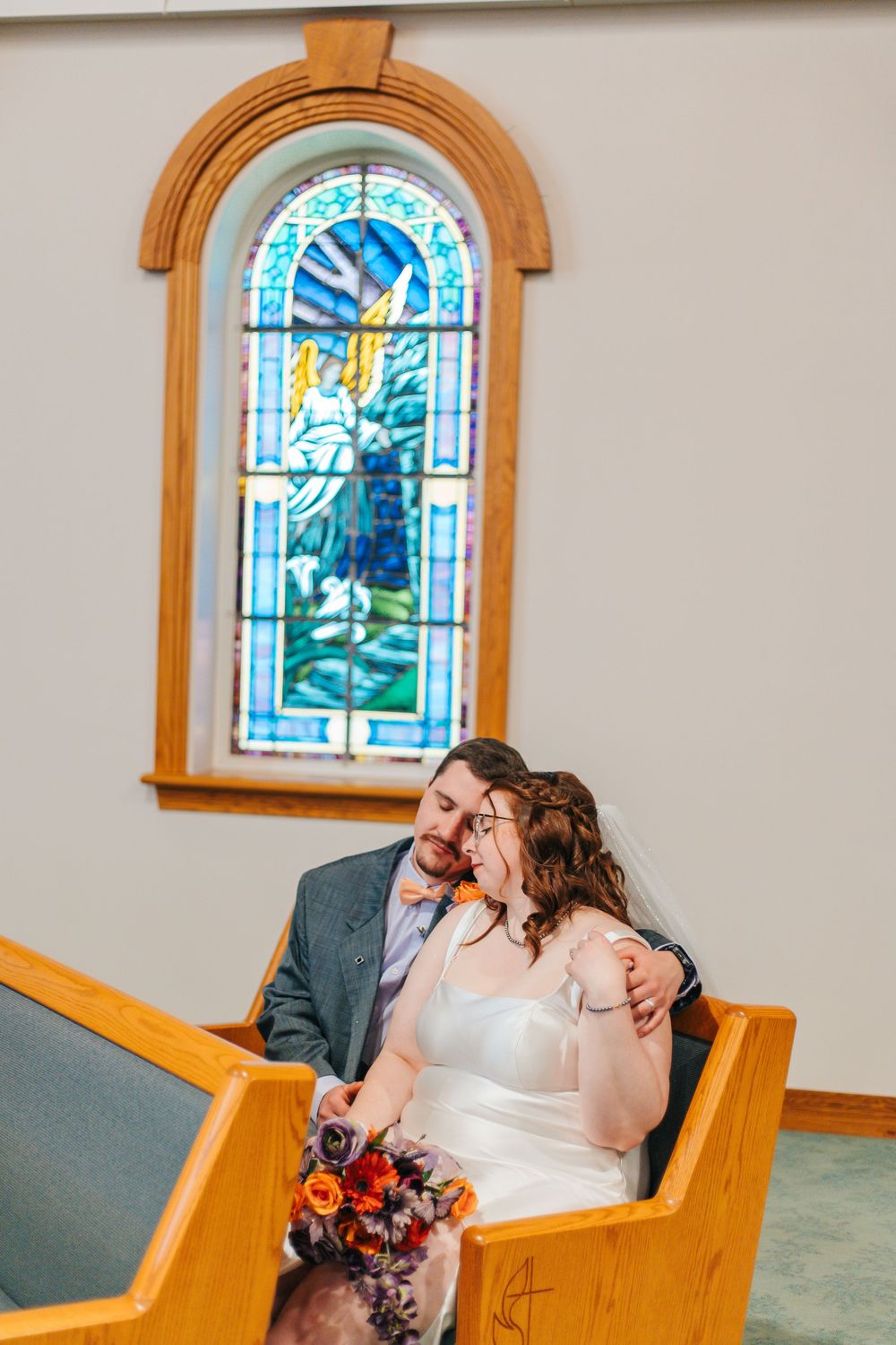 Bride and groom sit together in a church pew, smiling, with a colorful stained glass window behind them.