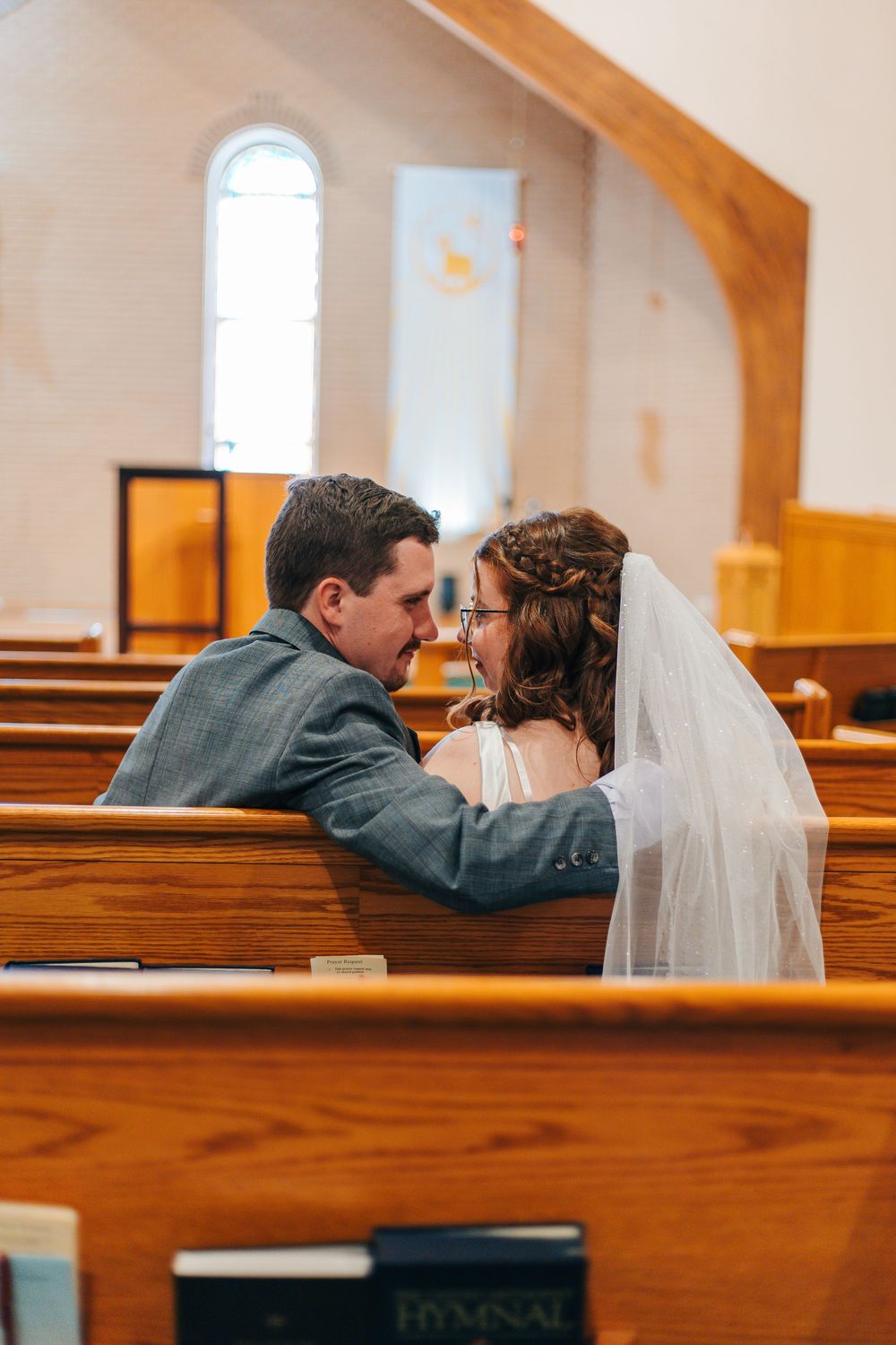 Newlywed couple sharing an intimate moment in wooden church pews, bride wearing veil and groom in gray suit.
