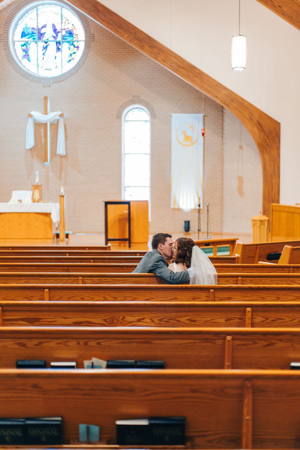Bride and groom share a kiss in wooden church pews with stained glass windows and altar in background.