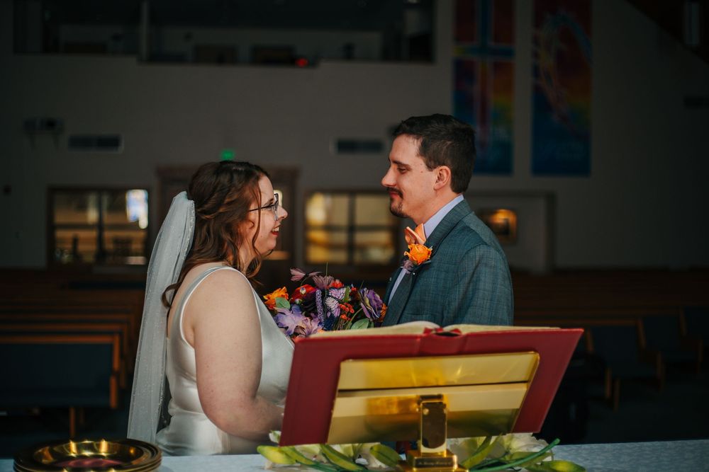 Bride and groom exchange vows at church altar, bride in white gown with veil, groom in gray suit with orange boutonniere.