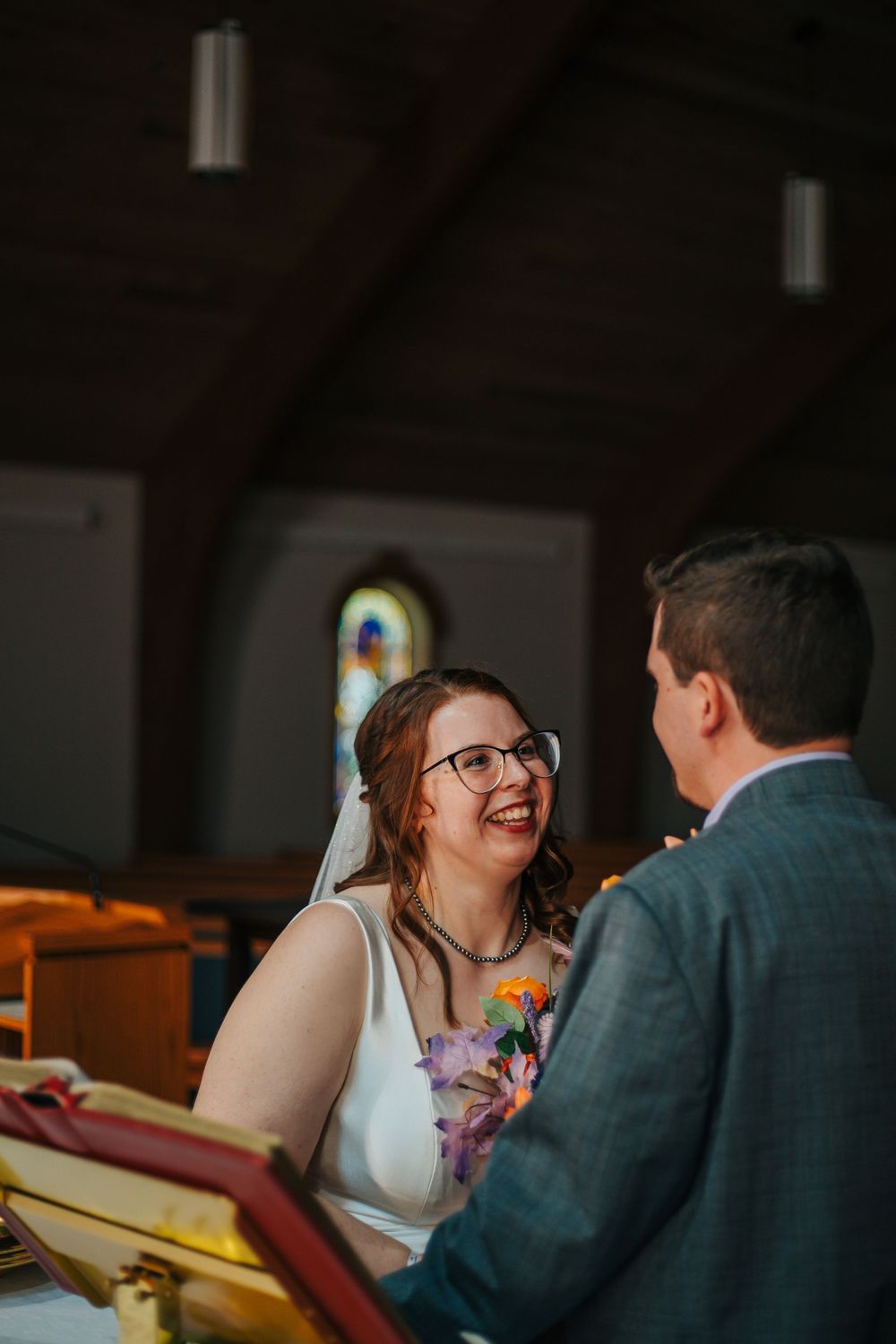Bride smiling joyfully at groom during wedding ceremony in a church with stained glass windows.