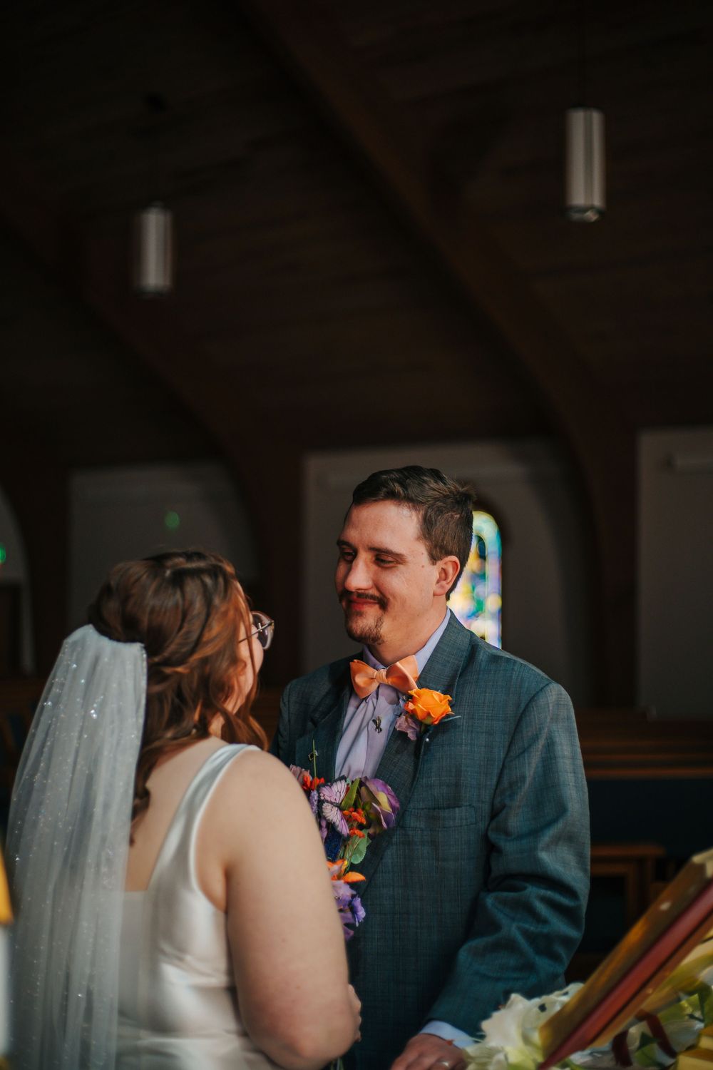 Groom smiles at bride during wedding ceremony inside a rustic wooden church with stained glass windows.