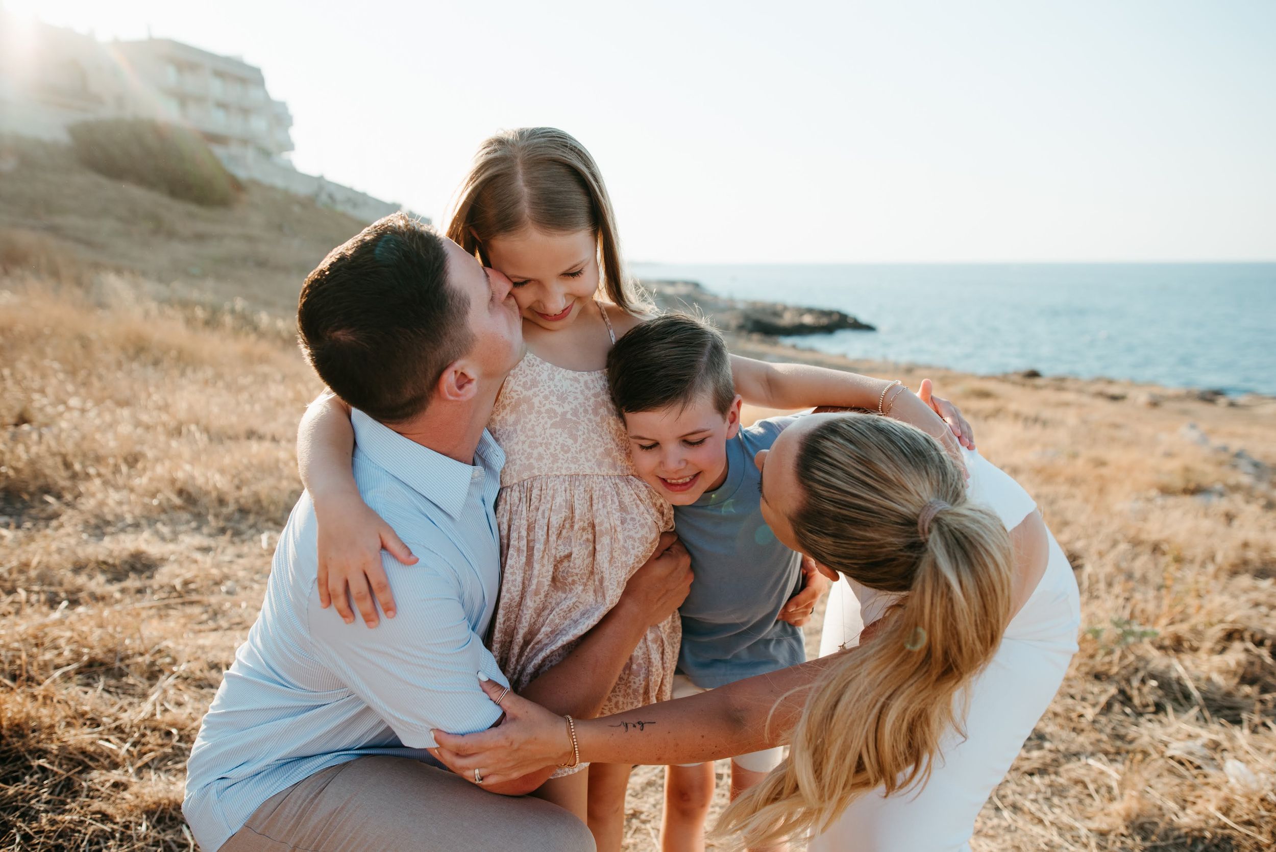 Happy family with two young children embracing and laughing together on a sunny coastal hillside.