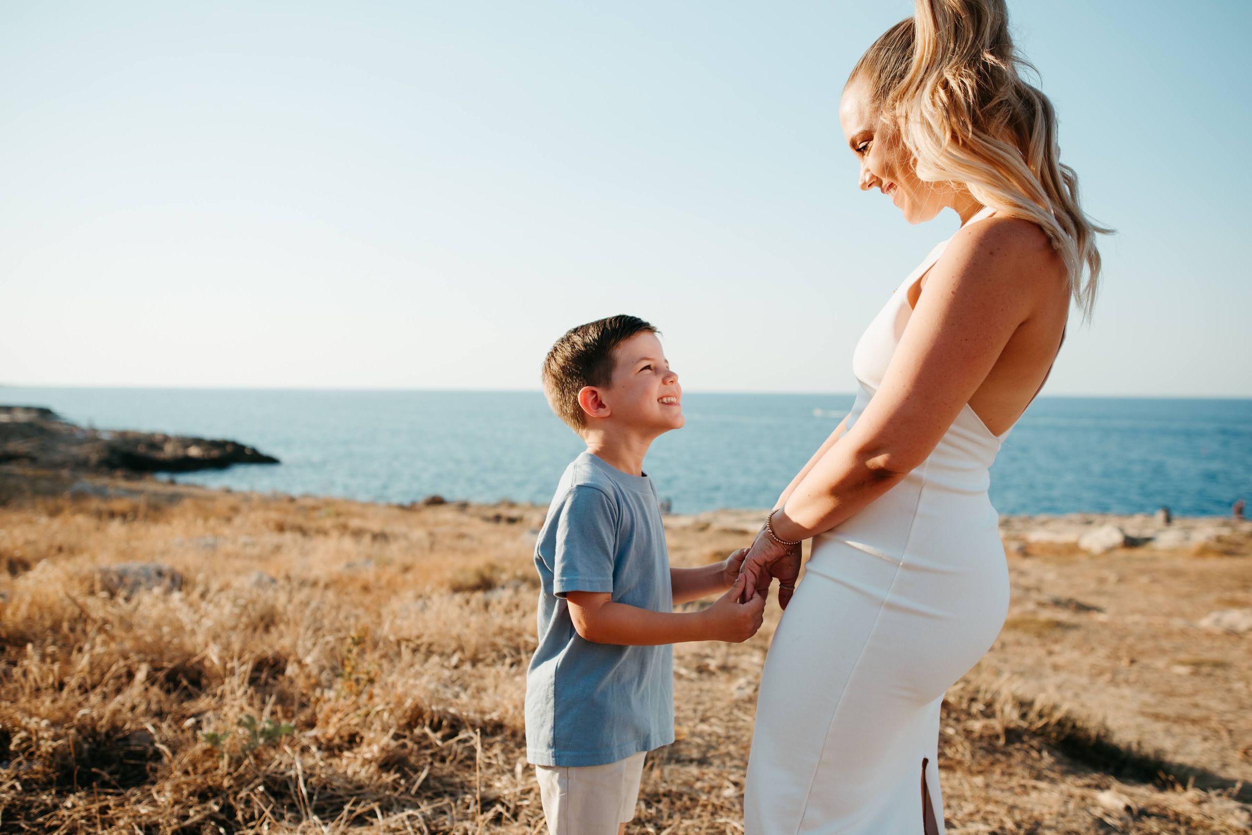 Pregnant woman in white dress holds hands with young boy by the sea on a sunny day.