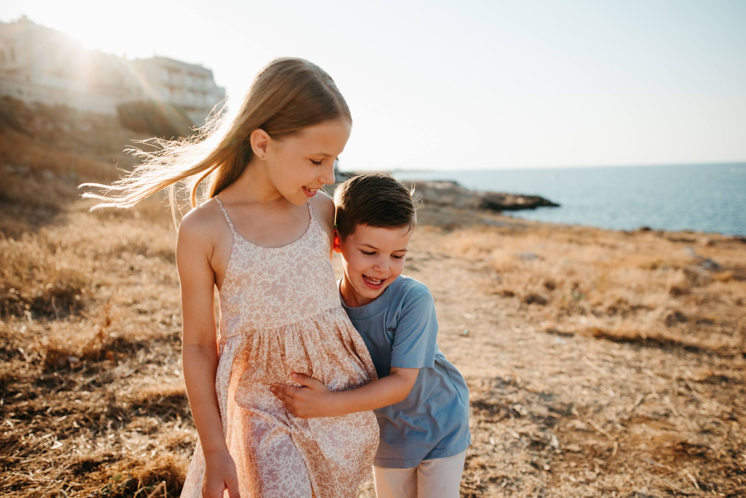 Two happy children hugging on a sun-drenched coastal hillside with the sea in the background.