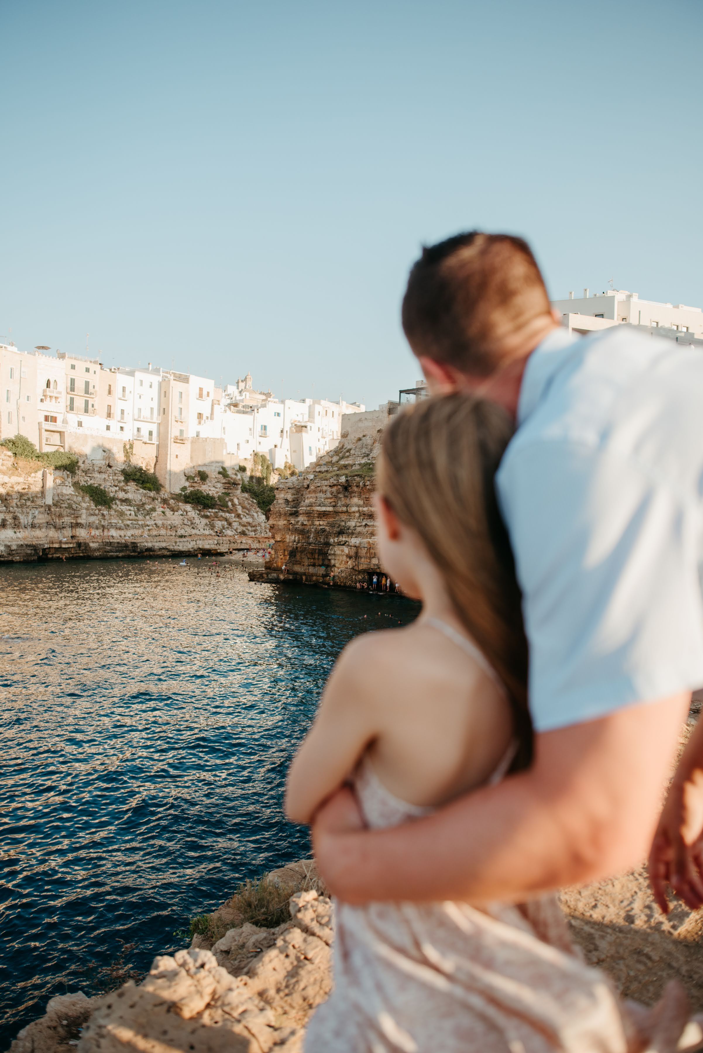 Romantic couple embracing on rocky cliffs overlooking a Mediterranean coastal town with blue waters.