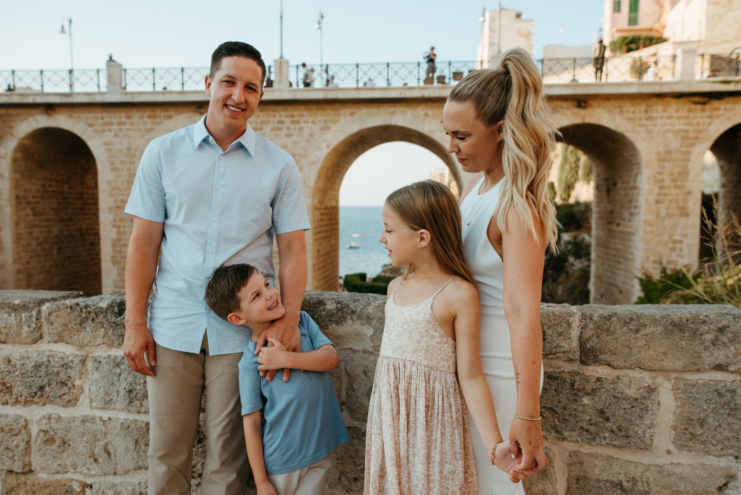 Family of four posing outdoors near stone arches with ocean views in background.
