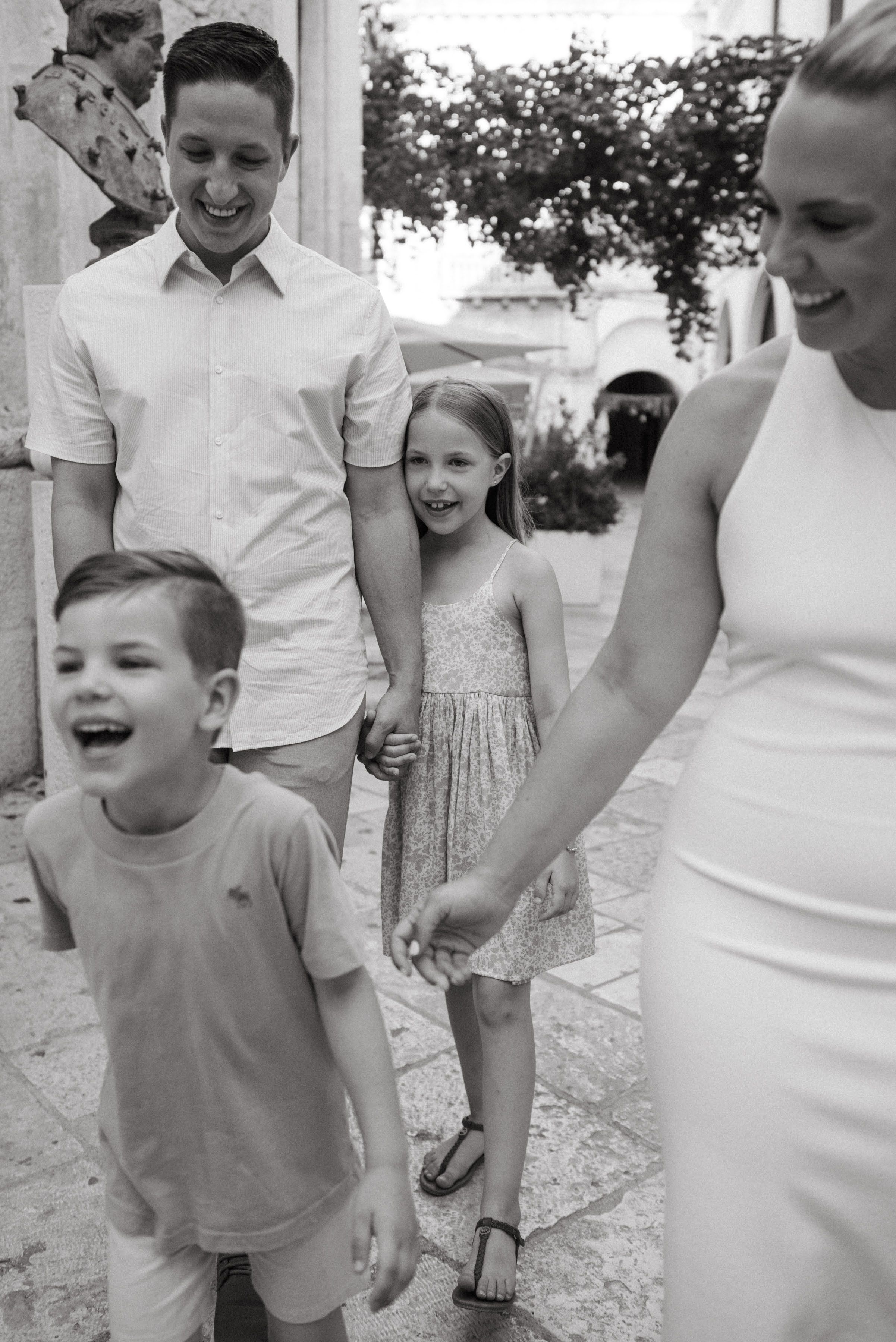 Black and white photo of a happy family with two children walking and laughing together on a sunny outdoor street.