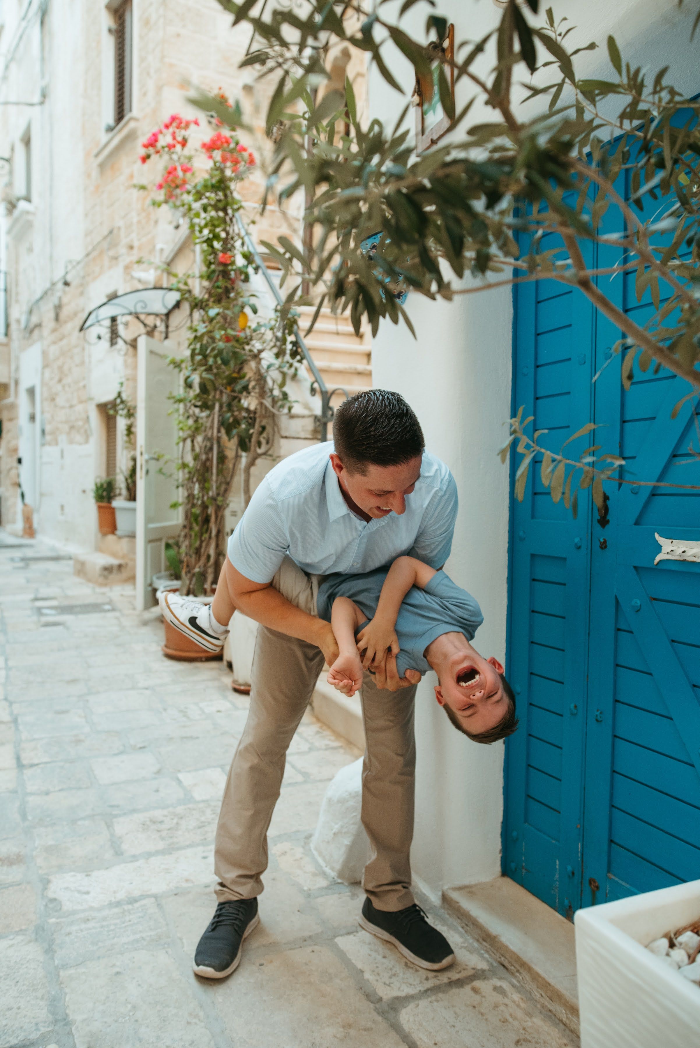 A man playfully lifts a laughing child upside down on a charming Mediterranean alley with blue door.