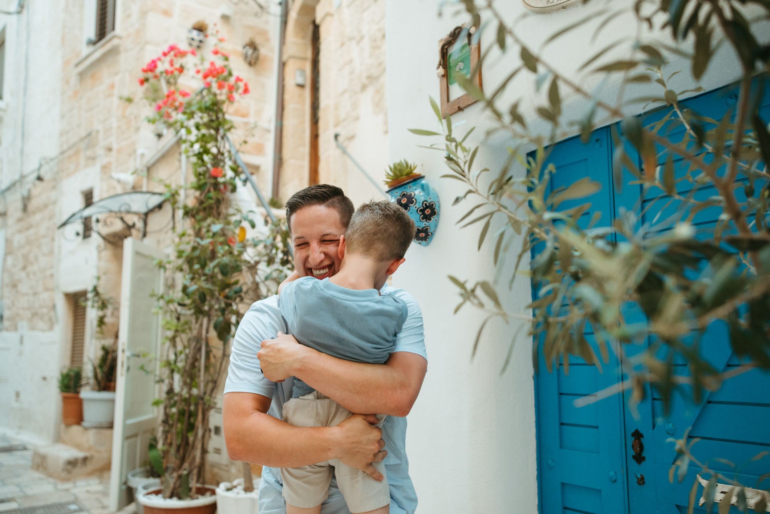 Father joyfully hugs young boy in a charming whitewashed Mediterranean alleyway.