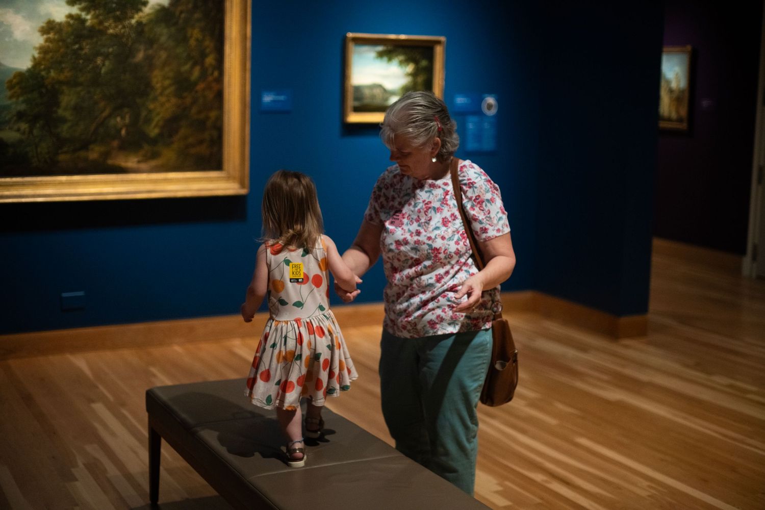 Woman and young girl interact in a blue-walled art museum gallery with landscape paintings on display.