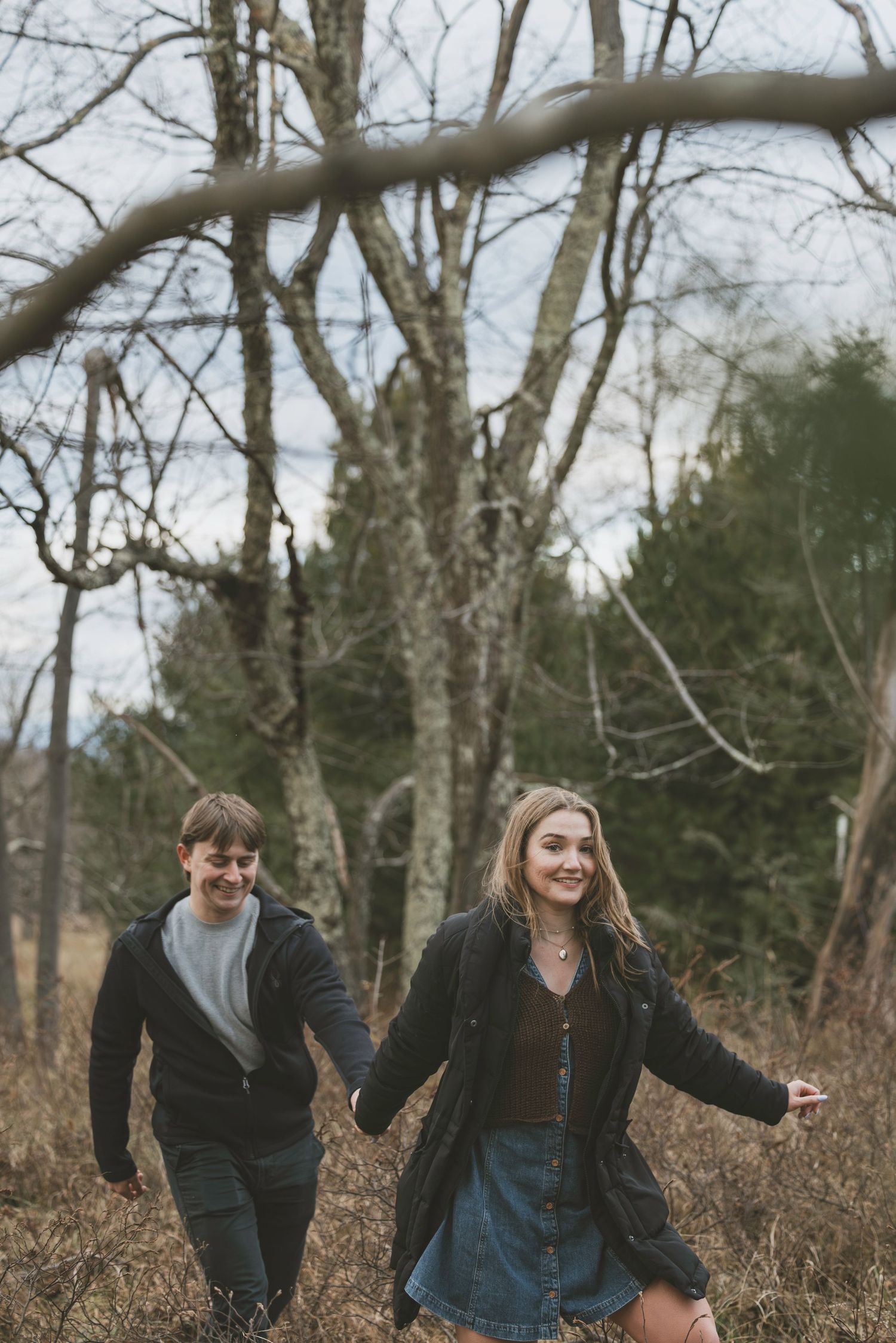 Young couple holding hands and walking through a wooded area in autumn, wearing casual jackets and smiling.