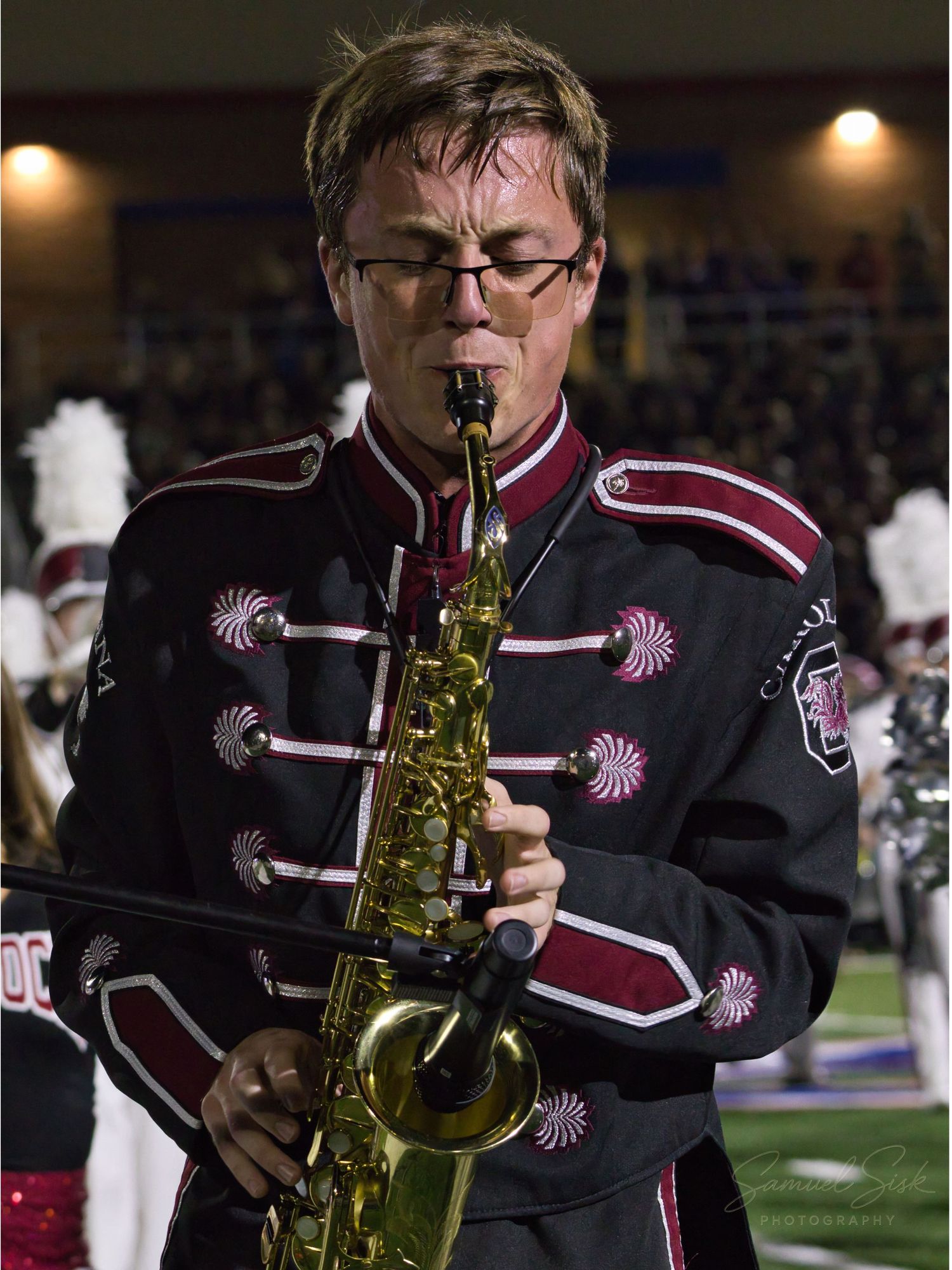 A marching band musician in a black and maroon uniform plays alto saxophone during a night football game.