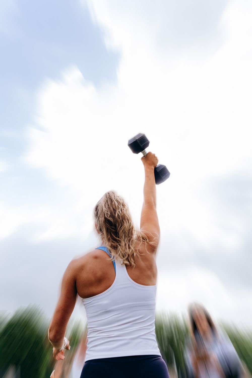 Woman in white tank top lifting a dumbbell overhead outdoors against a bright cloudy sky.