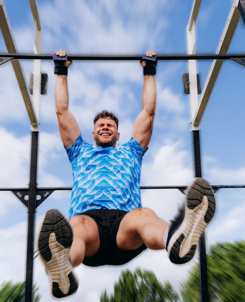 Muscular man in blue shirt doing pull-ups on outdoor bar against cloudy sky.
