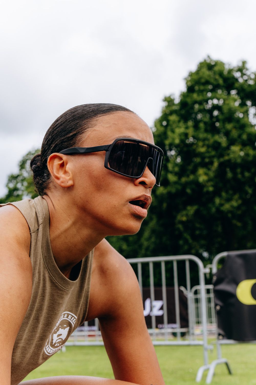 Athletic woman in sunglasses and olive tank top leaning forward outdoors at a sporting event.