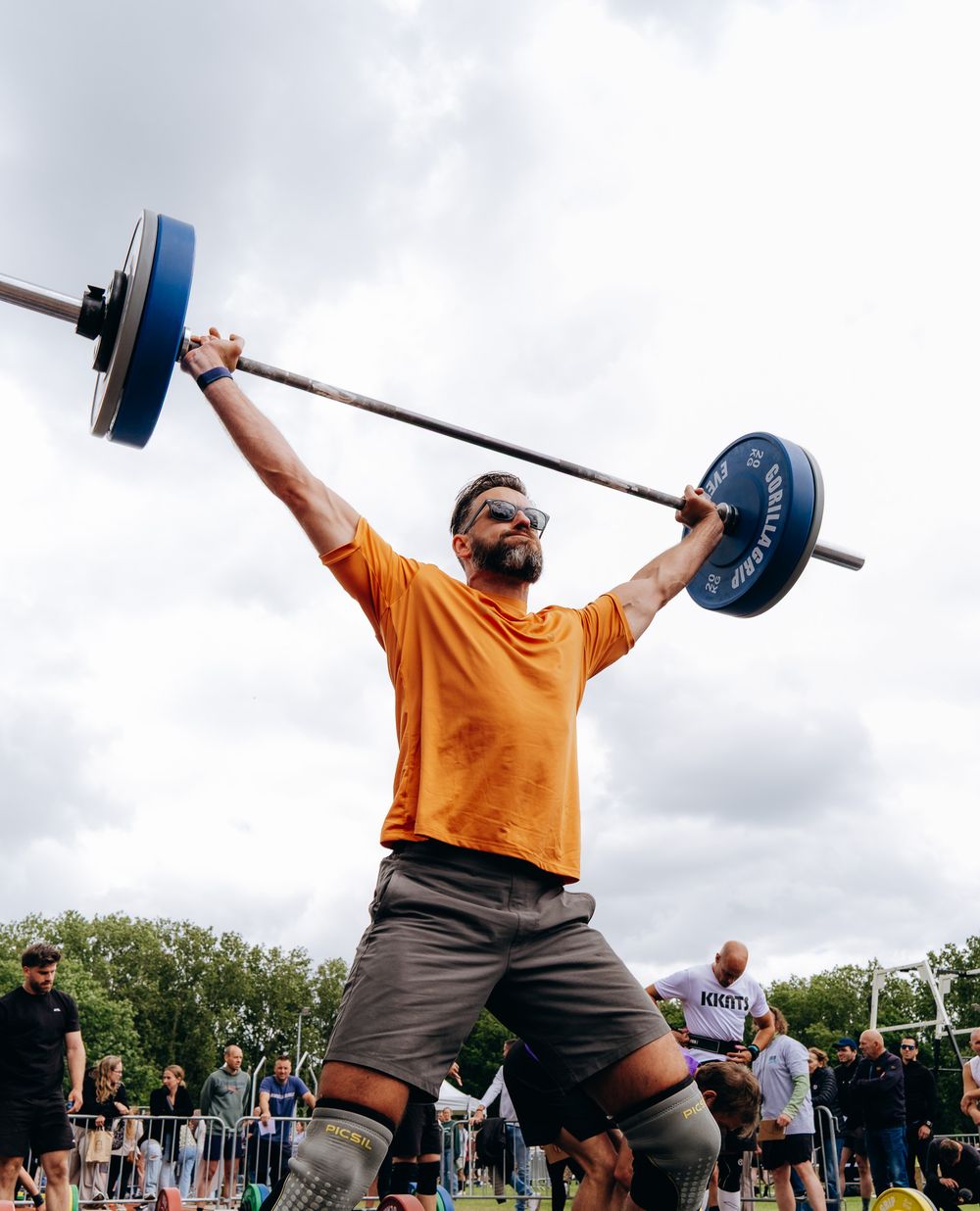 Athlete in orange shirt lifting heavy barbell overhead at outdoor competition.