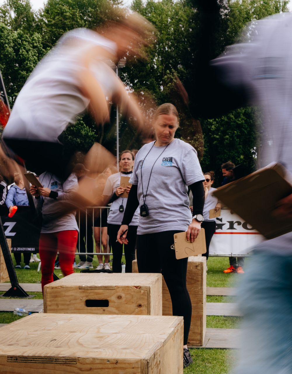 A judge oversees athletes box jumping at an outdoor fitness competition surrounded by blurred motion of competitors.