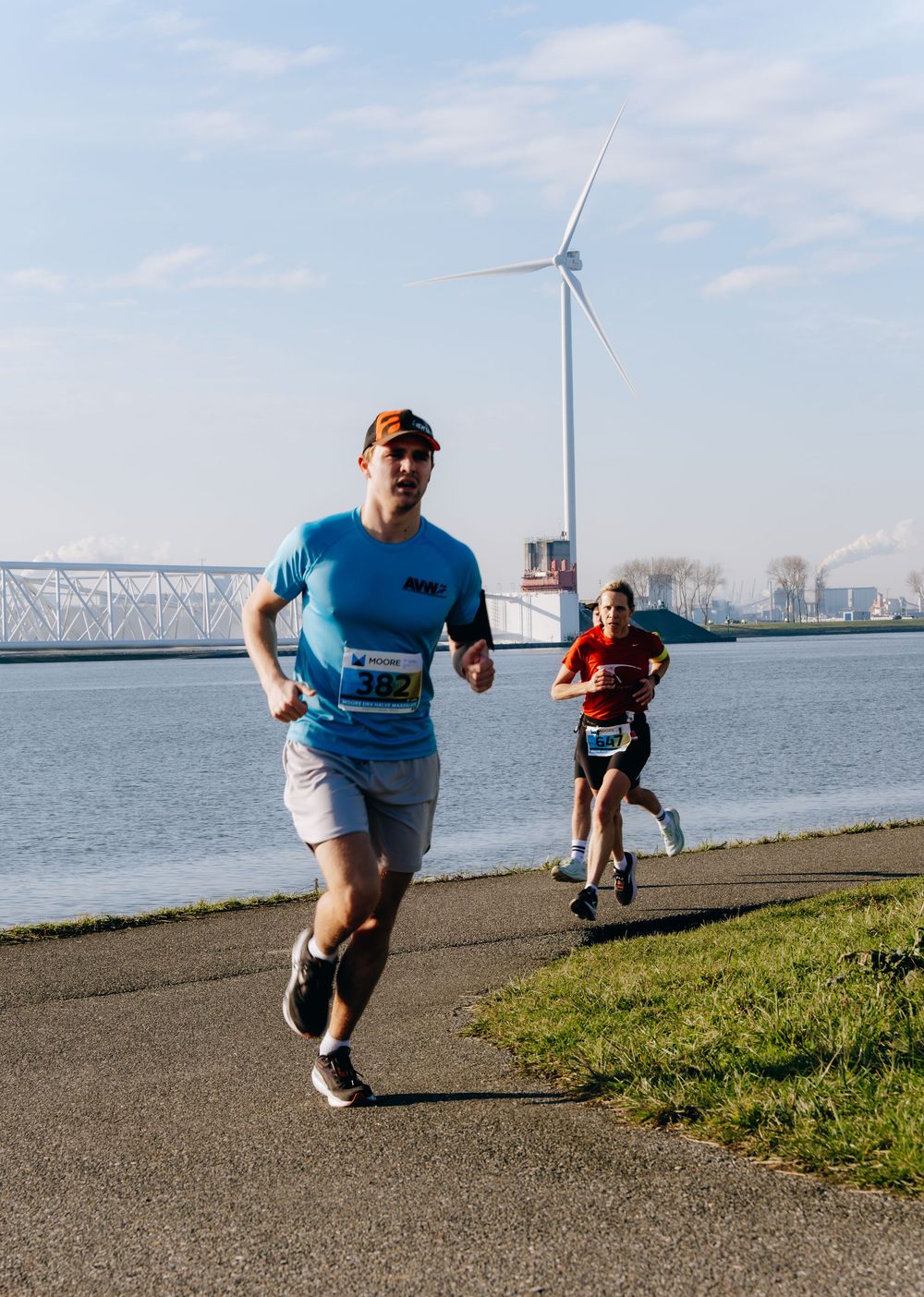 Two marathon runners racing along a waterfront path with a wind turbine and bridge in the background.