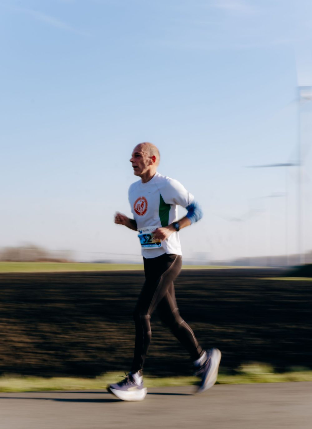 Bald male marathon runner in motion blur wearing a white race bib running under a clear blue sky.
