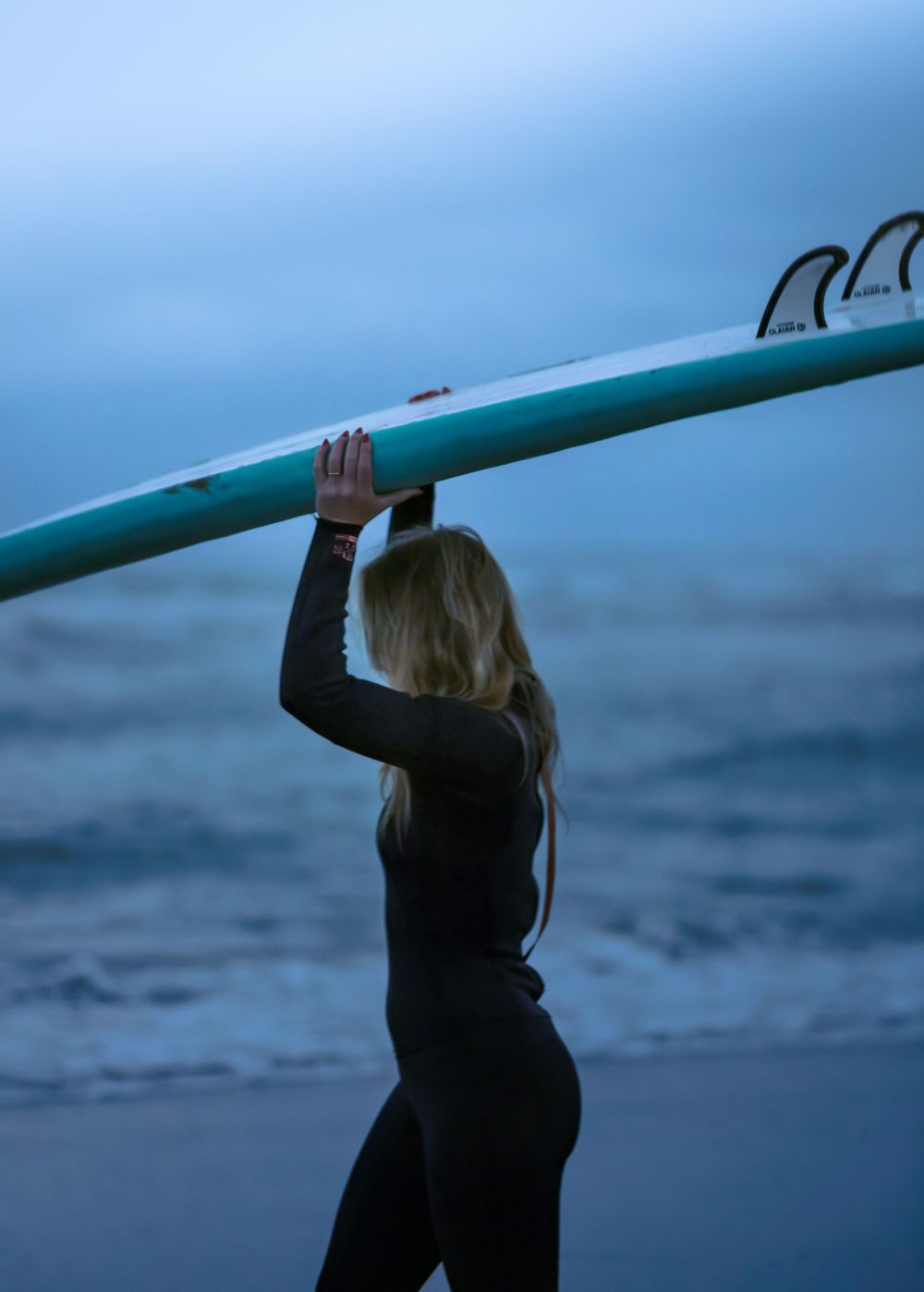 Blonde woman in wetsuit carrying teal surfboard on beach at dusk.