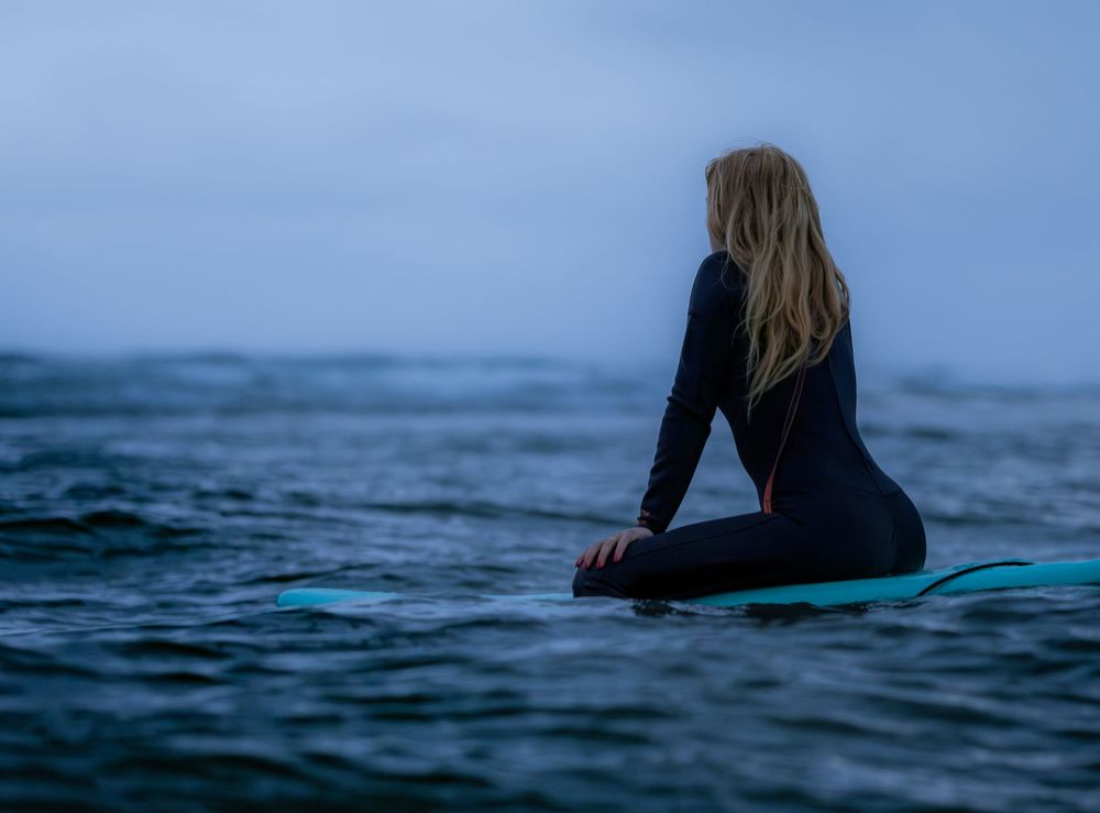 Female surfer sitting on teal surfboard in calm ocean at dusk.