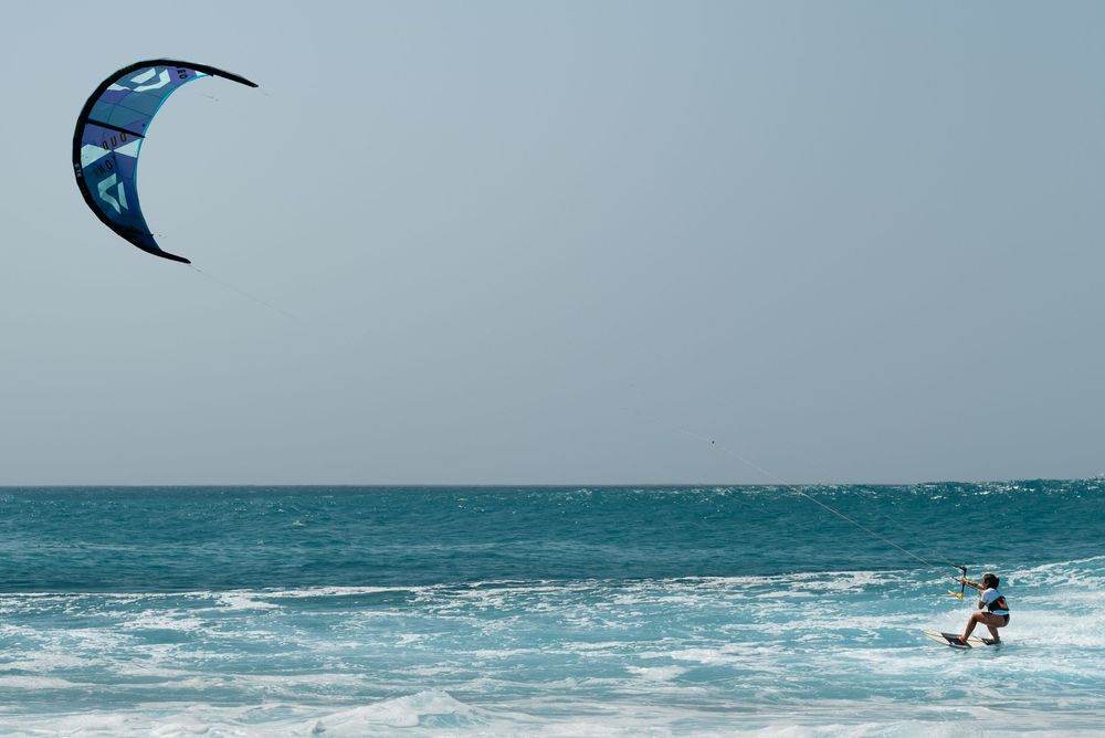 Kitesurfer riding ocean waves with blue kite against clear sky.