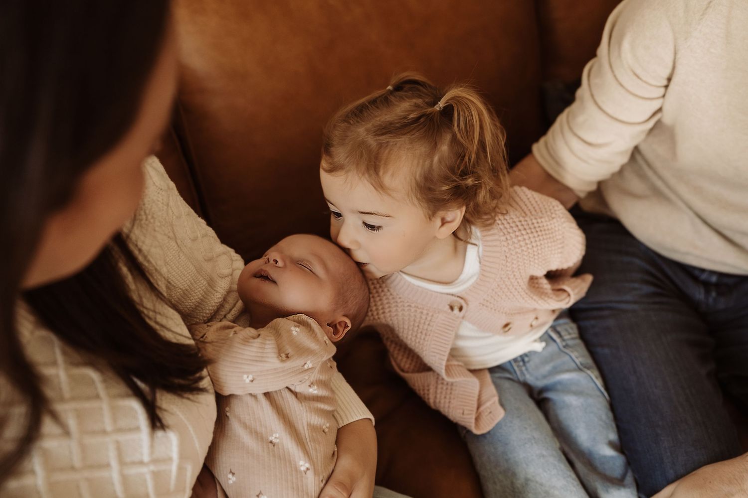 A toddler girl gently kisses a newborn baby held by their mother on a cozy brown armchair.