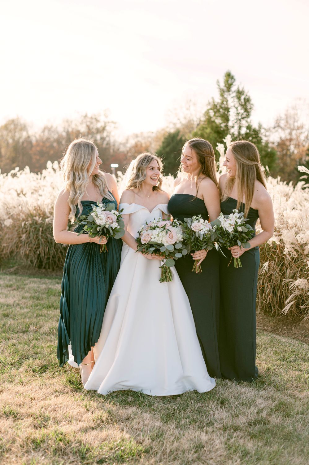 Bride laughing joyfully with three bridesmaids in dark green gowns holding bouquets at golden hour.