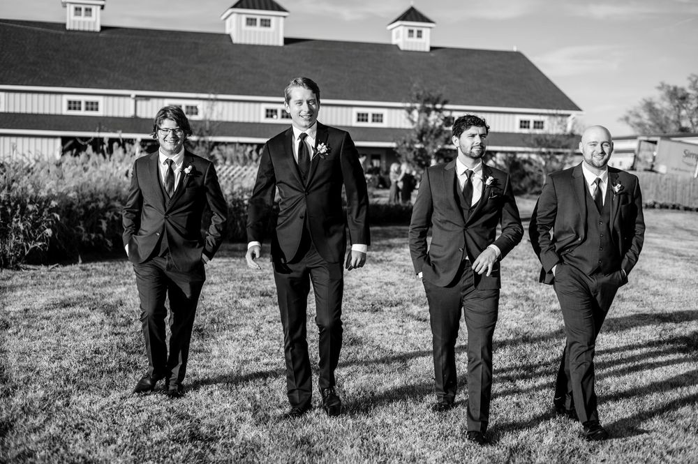 Black and white photo of groom and groomsmen in dark suits walking confidently outside rustic barn venue.