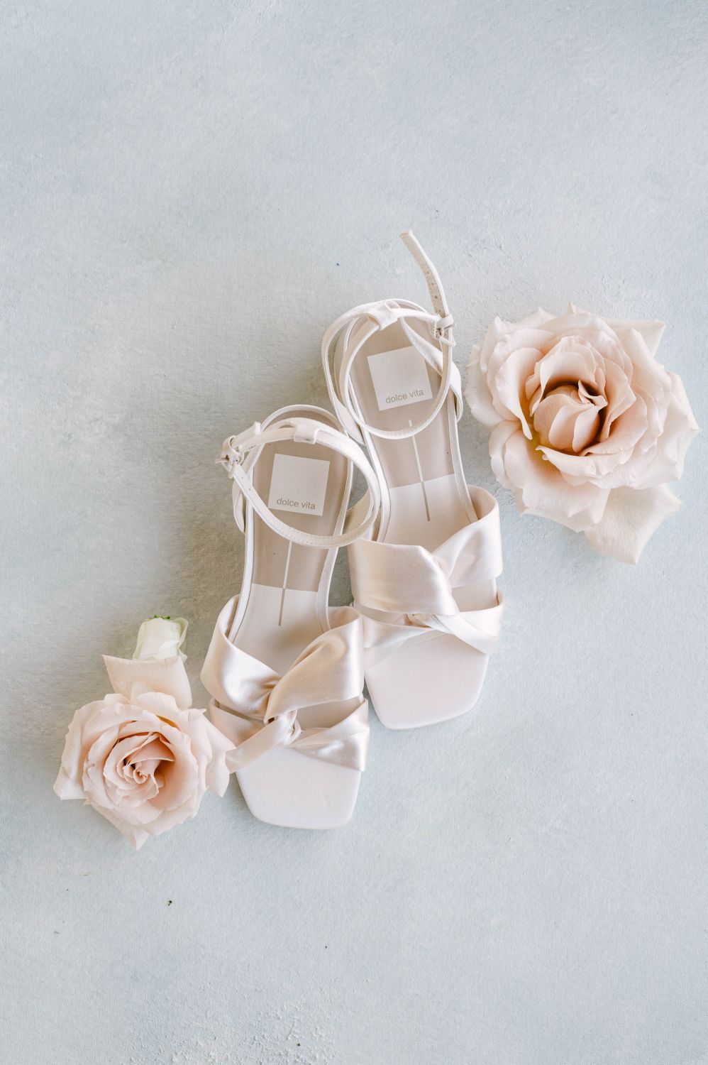 White satin bow wedding heels with ankle straps displayed alongside blush roses on a light background.