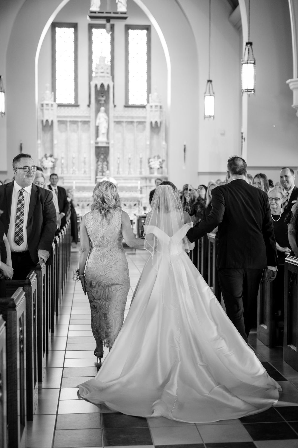 Black and white photo of bride walking down church aisle escorted by parents during traditional wedding ceremony.