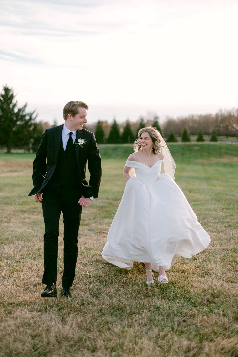 Bride in off-shoulder ball gown and groom in black suit walk hand in hand across a green field at golden hour.