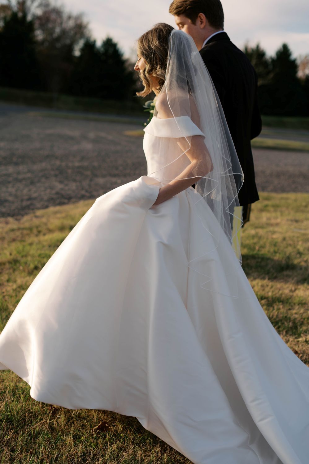 A bride in a flowing white ball gown and veil walks with a groom in a black tuxedo across a sunlit grassy field.