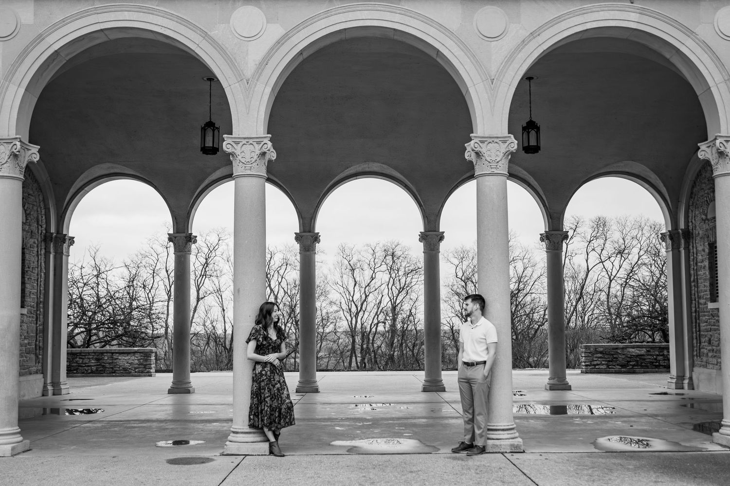 Couple stands apart under elegant stone arches in black and white engagement photo at a classical outdoor colonnade.