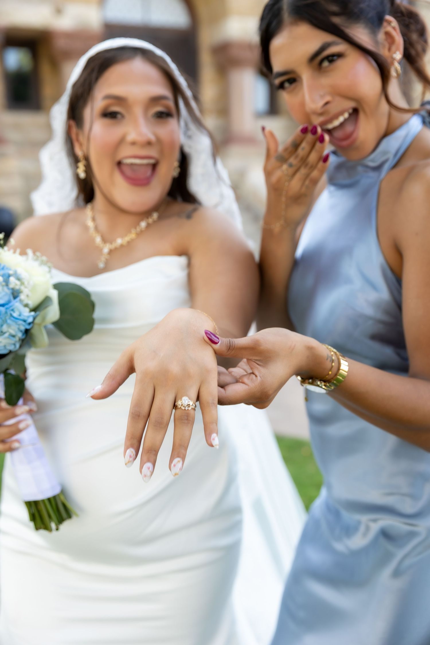 Bride and groom exchange rings during outdoor wedding ceremony with officiant in white robe and floral arch backdrop.