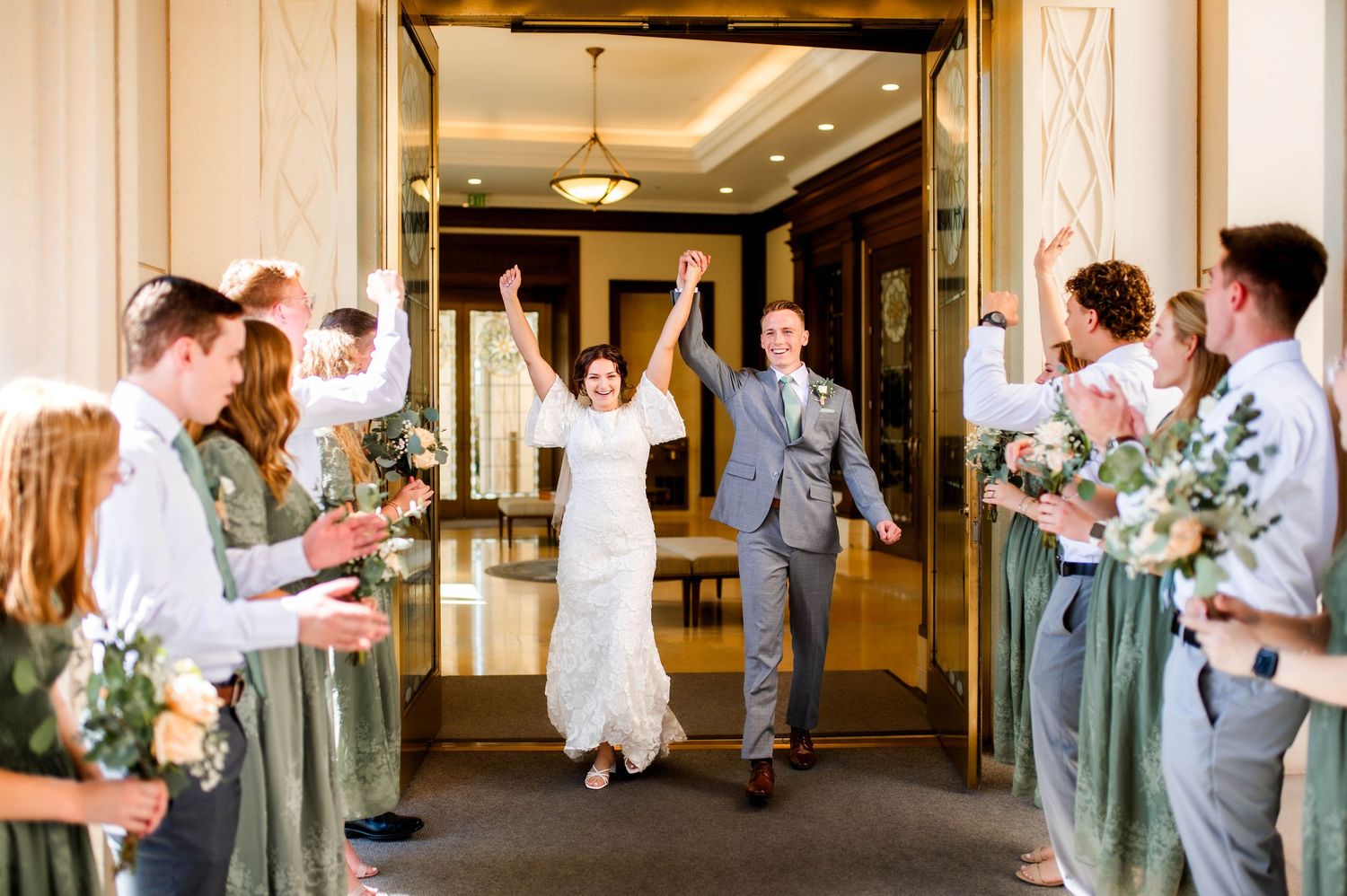 Joyful newlyweds exit a grand venue as wedding party cheers with bouquets lining the aisle.