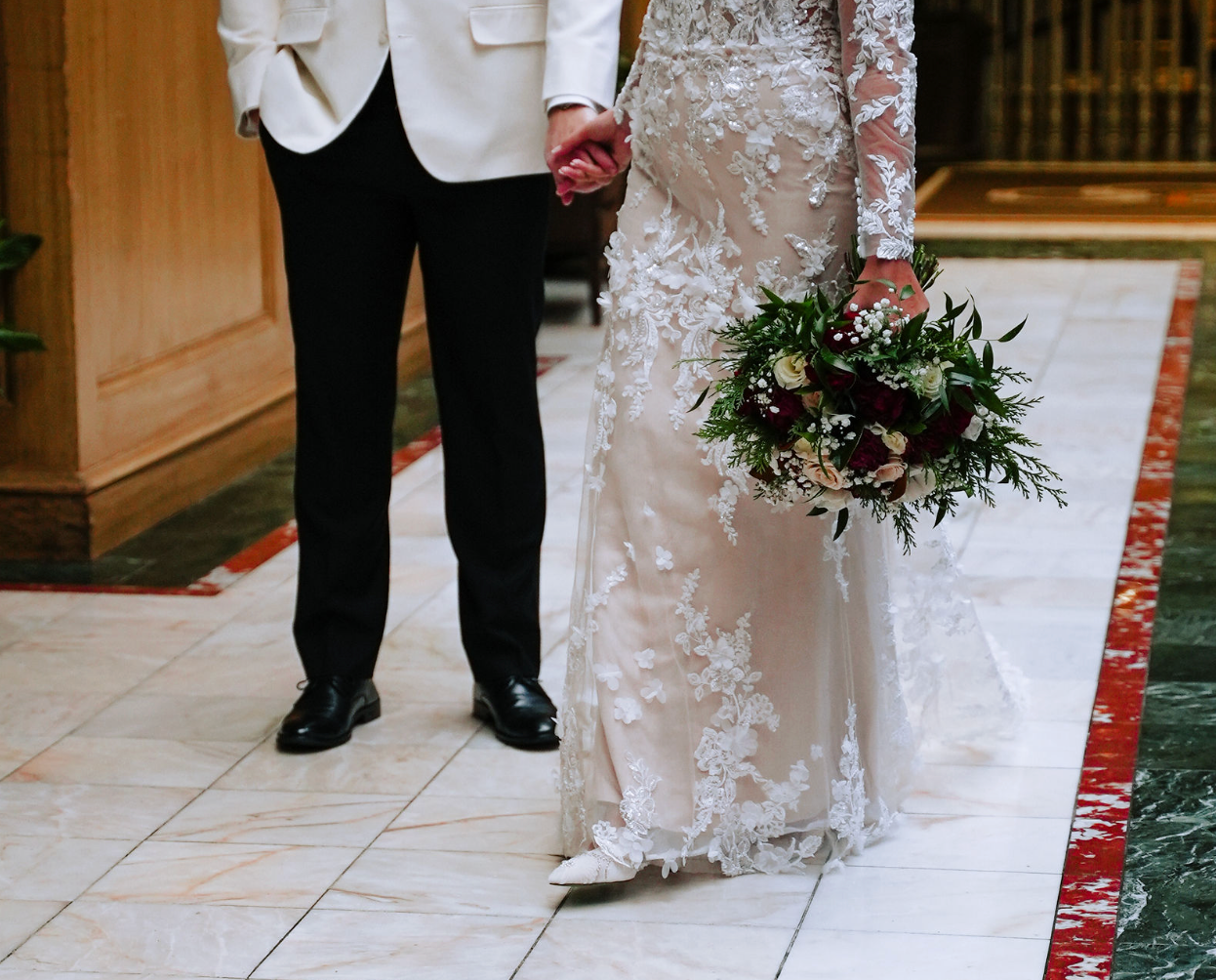 Bride in lace gown holding bouquet stands beside groom in white tuxedo jacket on marble floor.