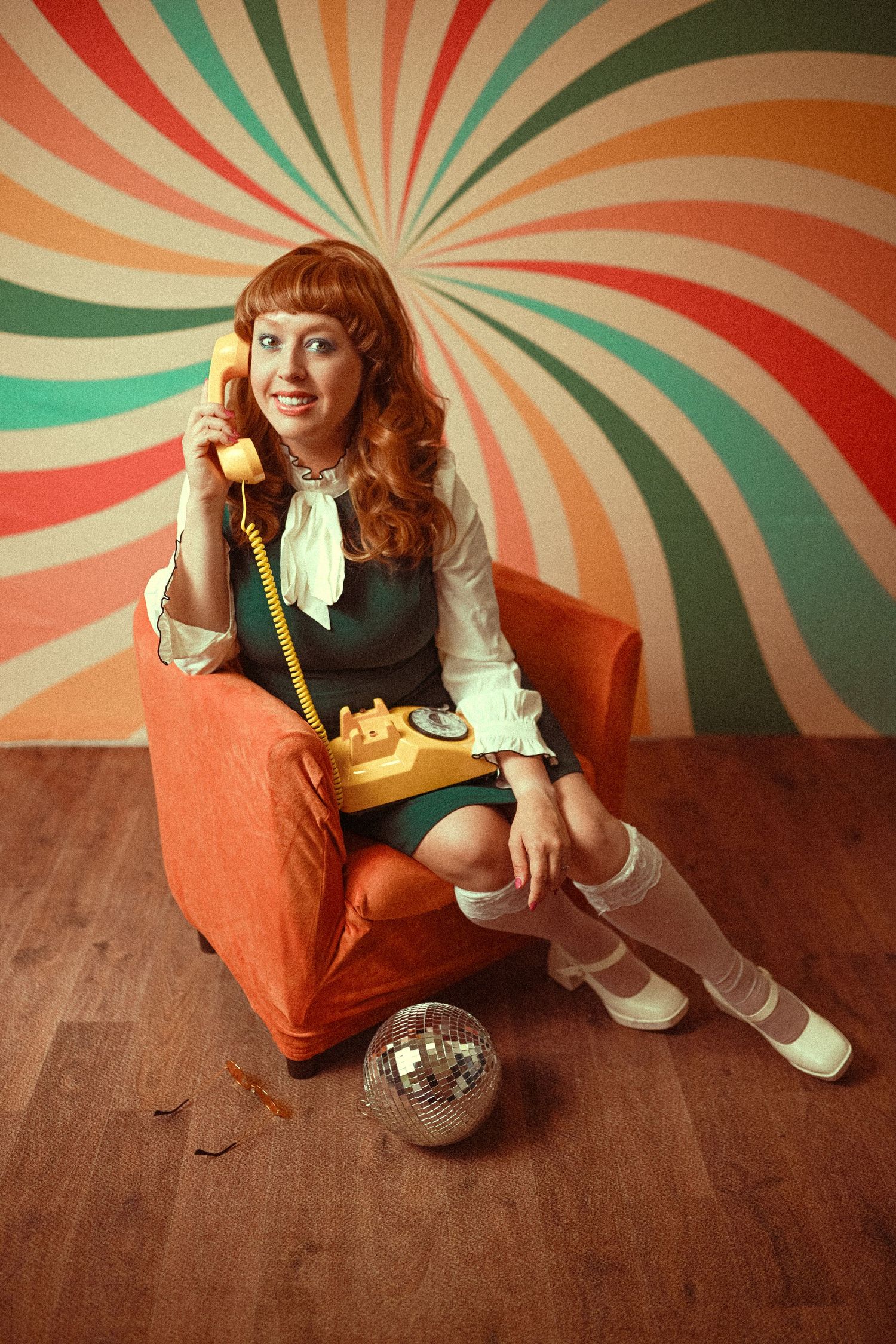 Woman in green dress sits in orange chair talking on yellow retro phone, disco ball nearby, colorful swirl backdrop.