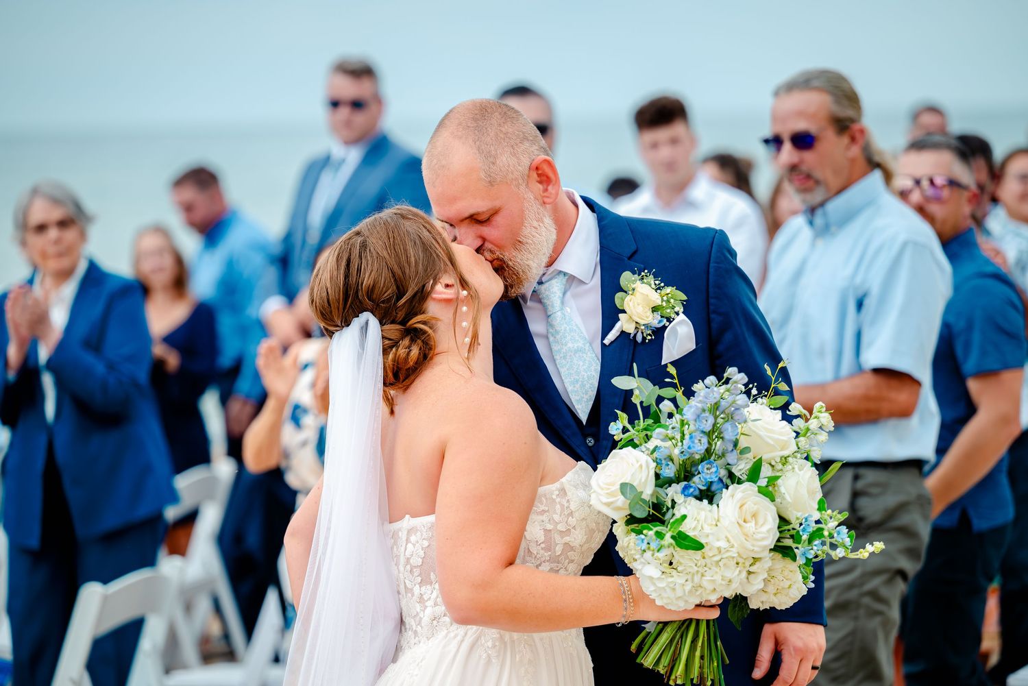 Bride and groom share first kiss at outdoor beach wedding ceremony with guests applauding in background.