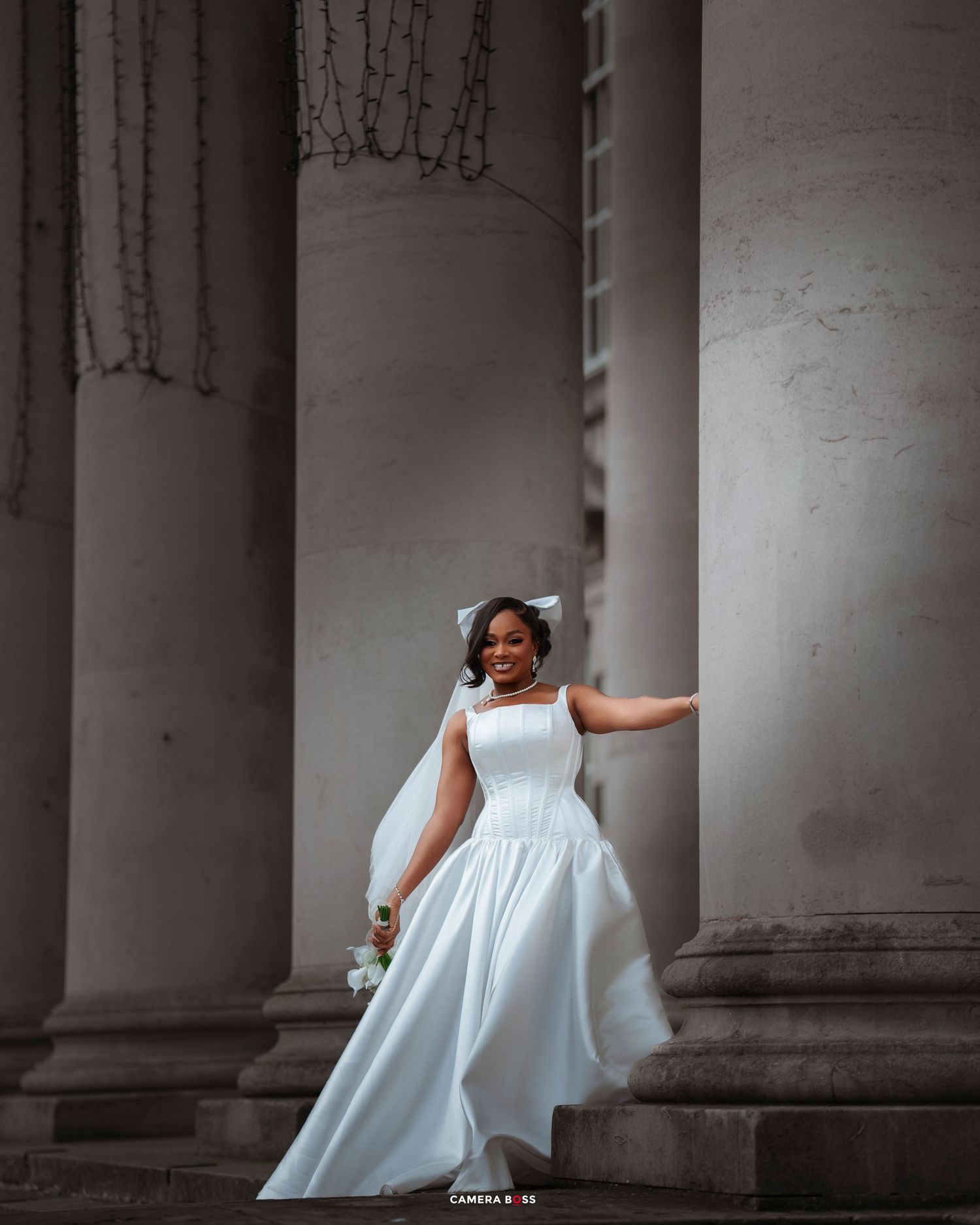 Bride in elegant white gown posing gracefully between grand stone columns, holding a bouquet.