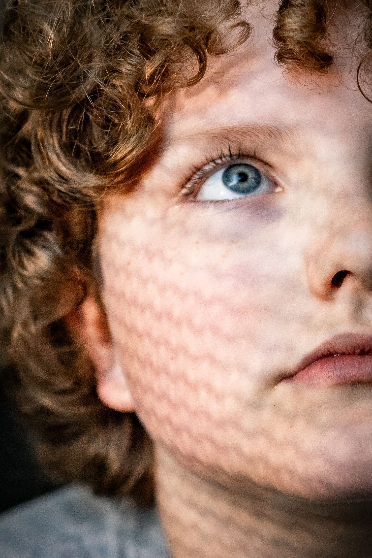 Close-up portrait of a curly red-haired child with blue eyes and freckles, gazing upward thoughtfully.