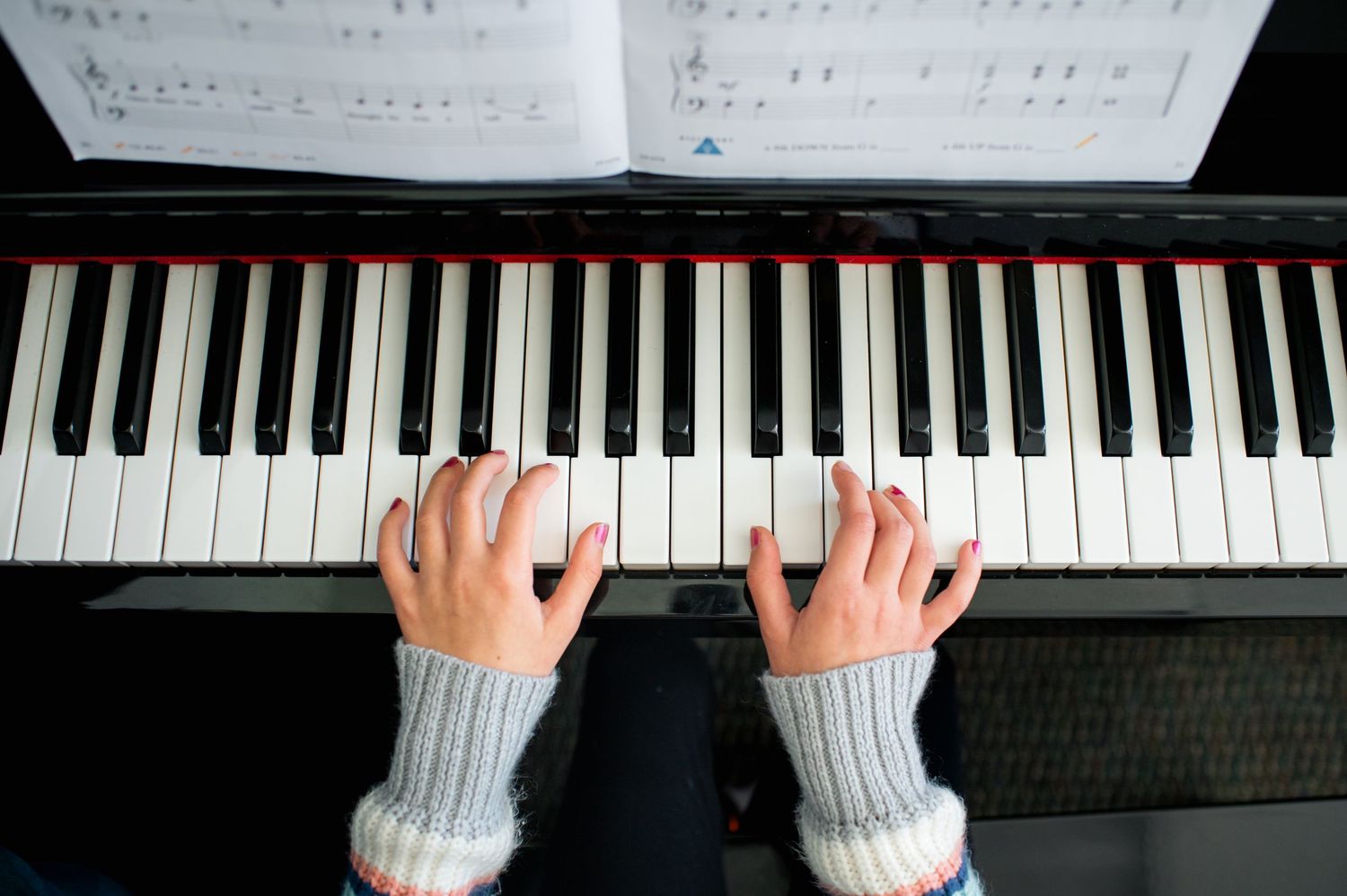Overhead view of hands playing piano keys with sheet music, wearing cozy sweater sleeves during a music lesson.