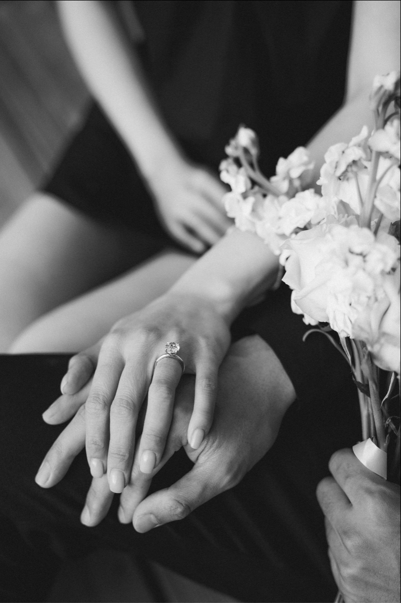 Black and white photo of couple holding hands with engagement ring and wedding bouquet of white flowers.