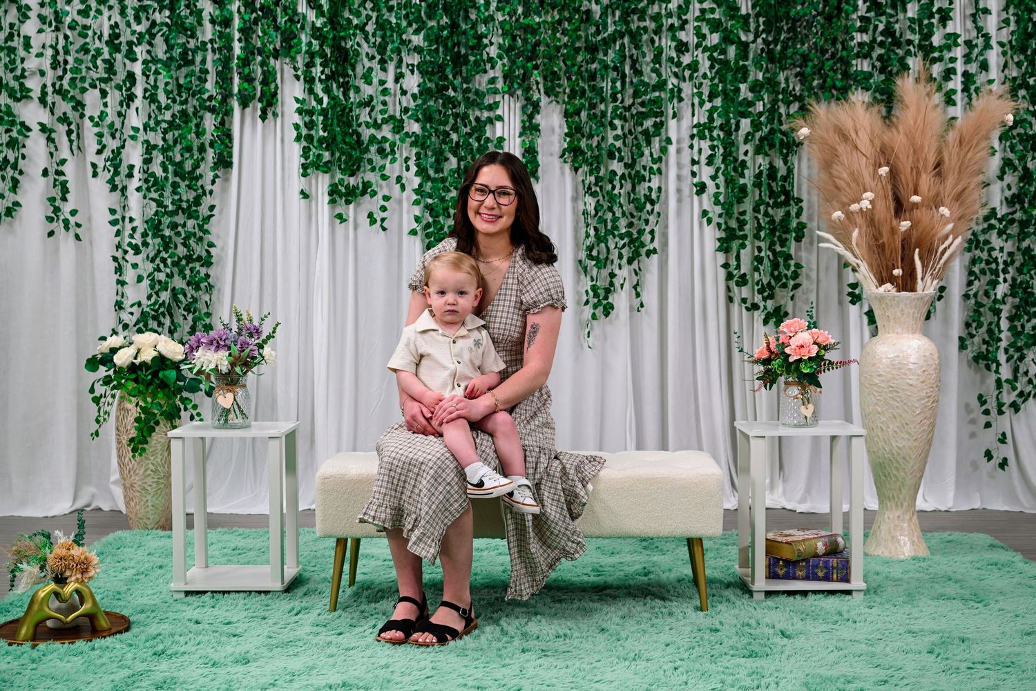 Woman in floral dress sitting on bench with toddler in decorated studio with green ivy backdrop.