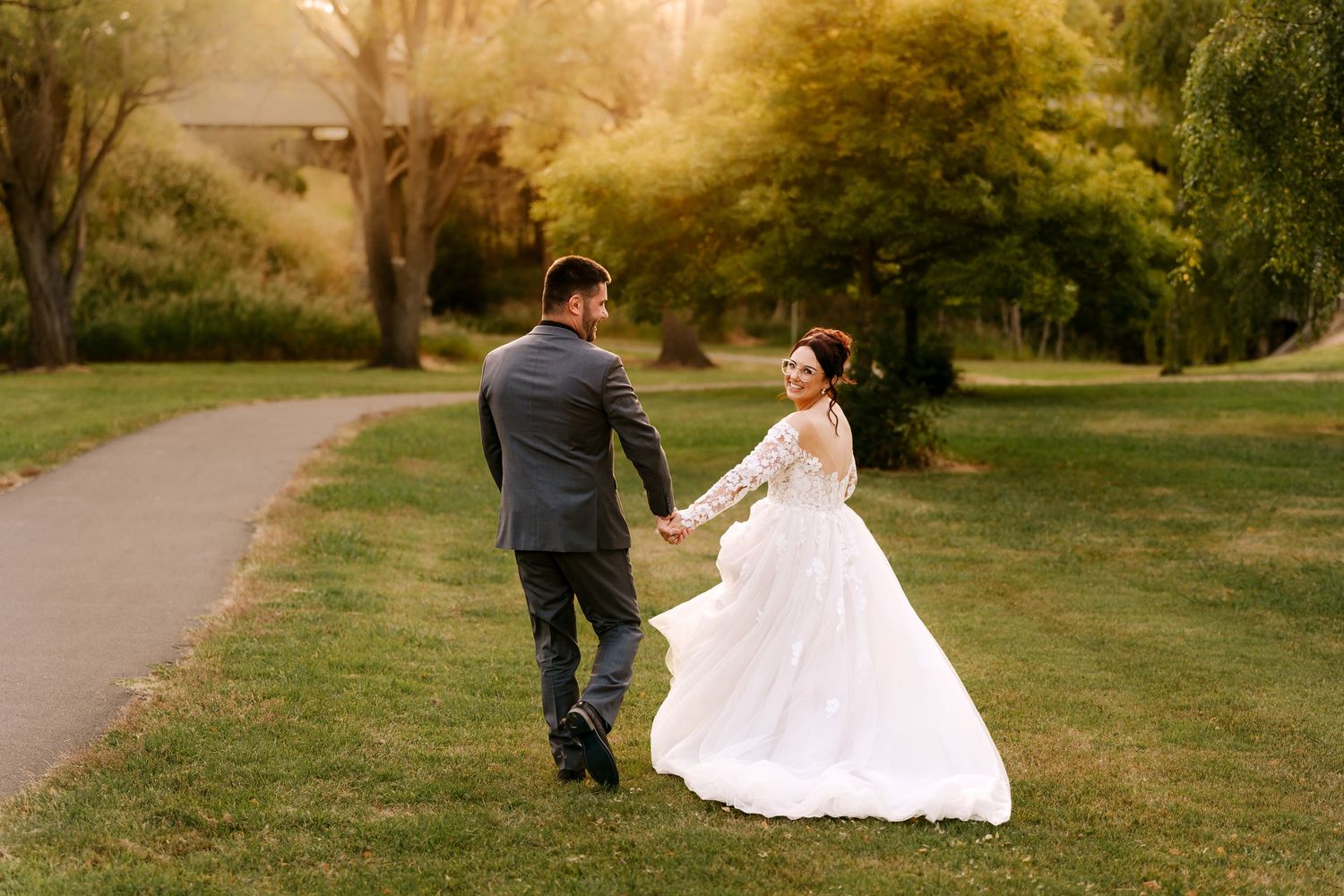 Bride and groom walk hand-in-hand through a sunlit garden, bride in lace gown, groom in gray suit.