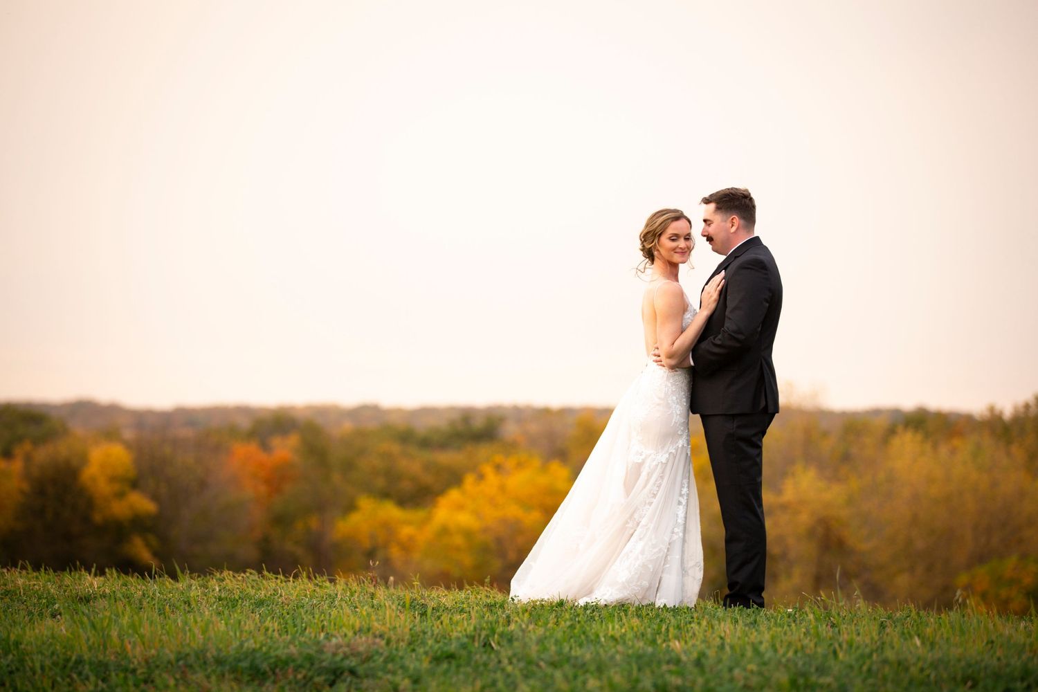 A bride and groom embrace on a hilltop overlooking an autumn landscape at sunset.  Iowa City wedding photographer.