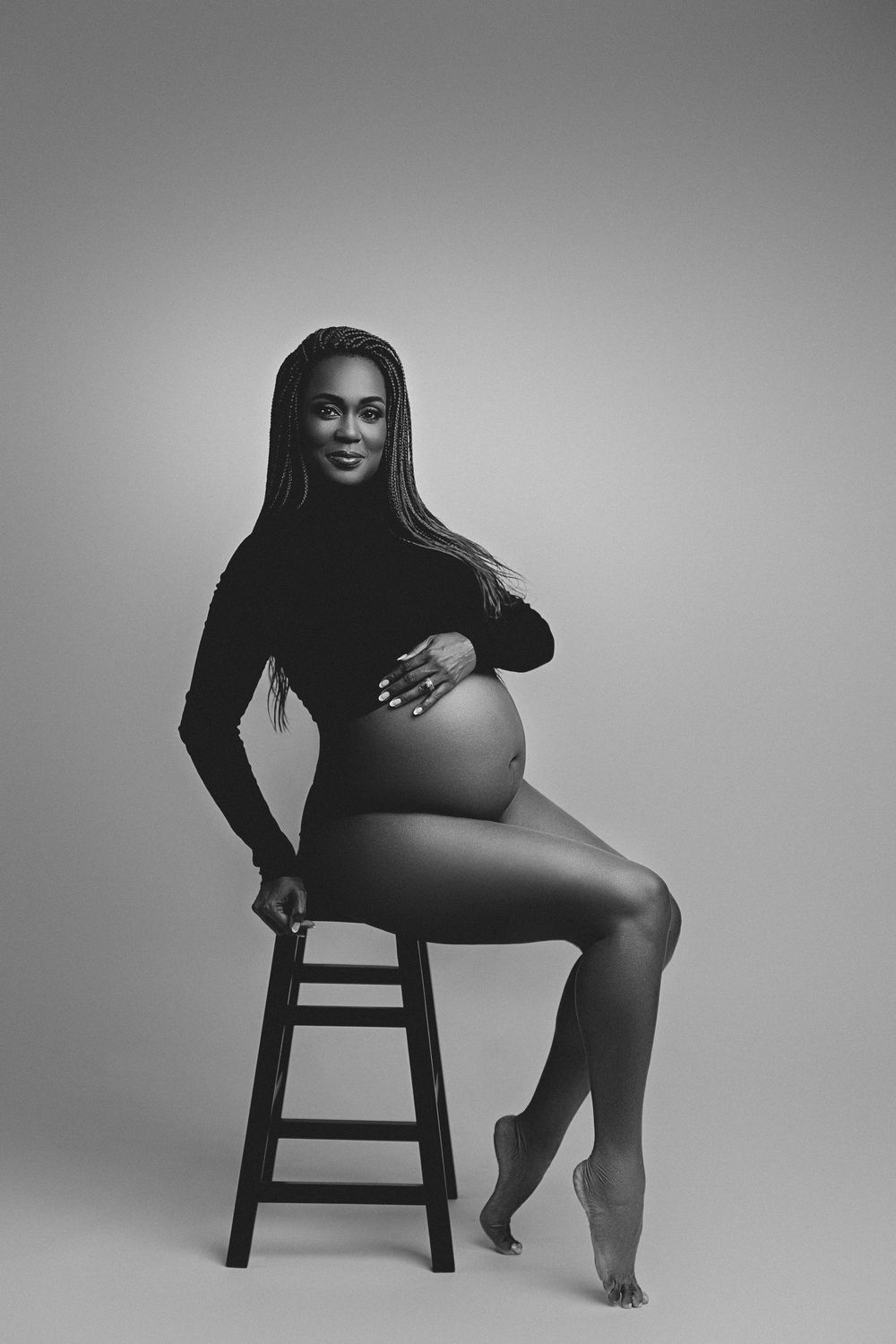 Black and white maternity photo of a pregnant woman sitting on a stool, cradling her baby bump in a studio setting.
