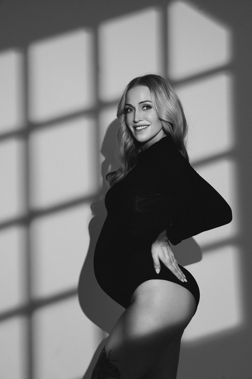 Black and white photo of a smiling pregnant woman in a black bodysuit posing gracefully with window light shadows.