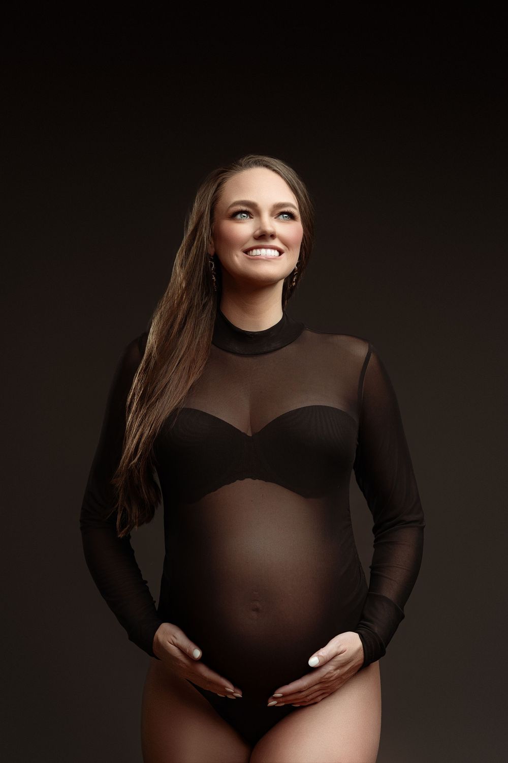 Smiling pregnant woman in black sheer bodysuit cradling baby bump against dark studio background.